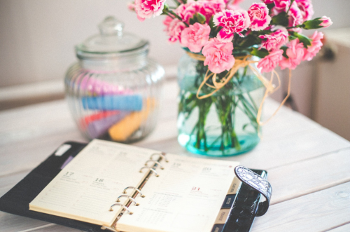 "Imagem de uma mesa elegantemente arrumada com um vaso de flores coloridas e uma agenda aberta. A mesa está organizada, refletindo a importância do planejamento para uma vida equilibrada. A agenda destaca a integração dos exercícios físicos diários na rotina, simbolizando o compromisso com o bem-estar e a saúde. O vaso de flores representa a vitalidade e a alegria que uma prática regular de atividades físicas pode trazer à vida cotidiana."